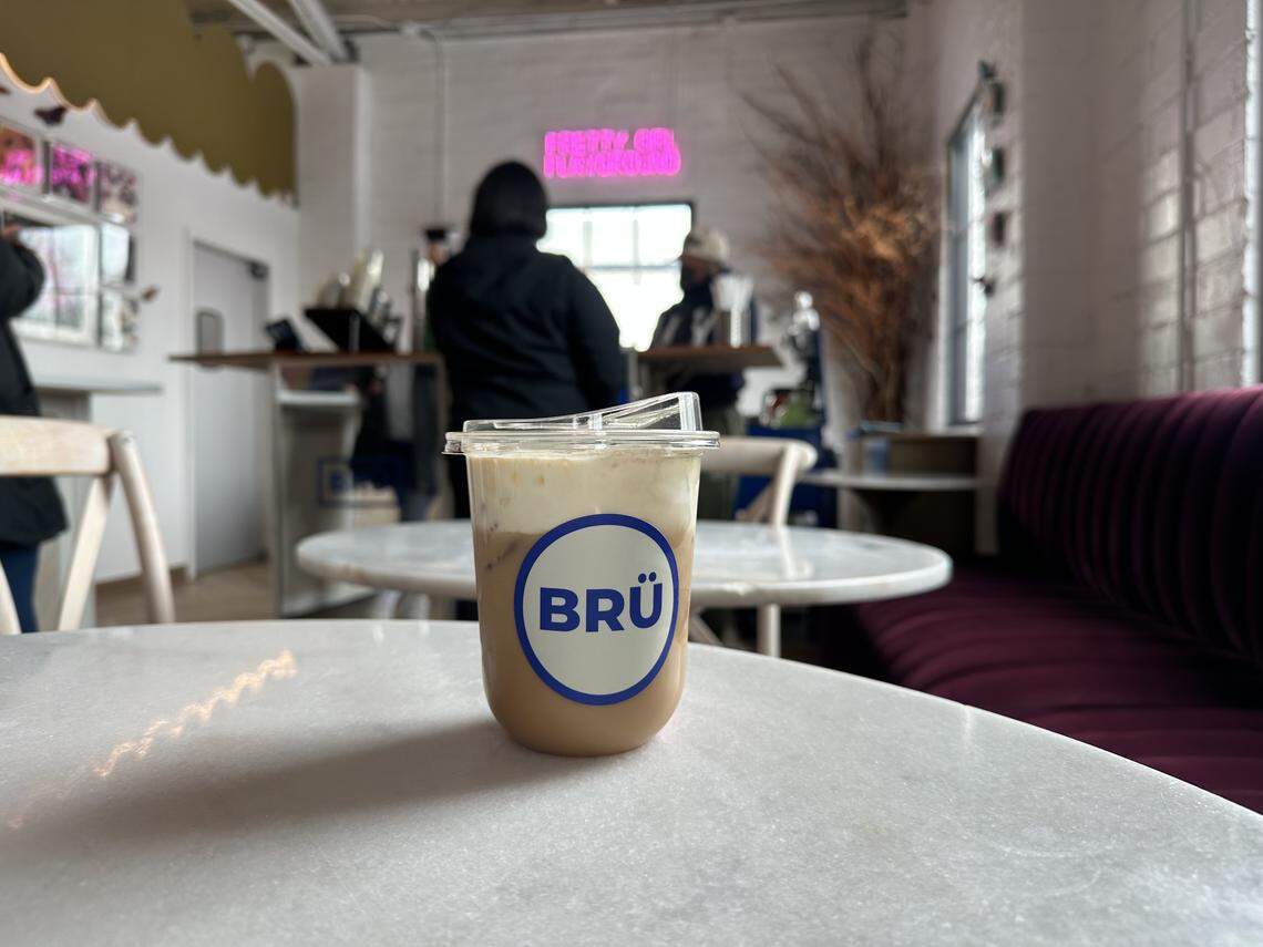 A close-up, focused shot of a BRÜ iced coffee drink sitting on a white marble table. The drink has a layered look with a thick layer of creamy foam on top. In the blurred background, the coffee shop interior is visible, including the “PRETTY GIRL PLAYGROUND” neon sign and the baristas working at the counter.