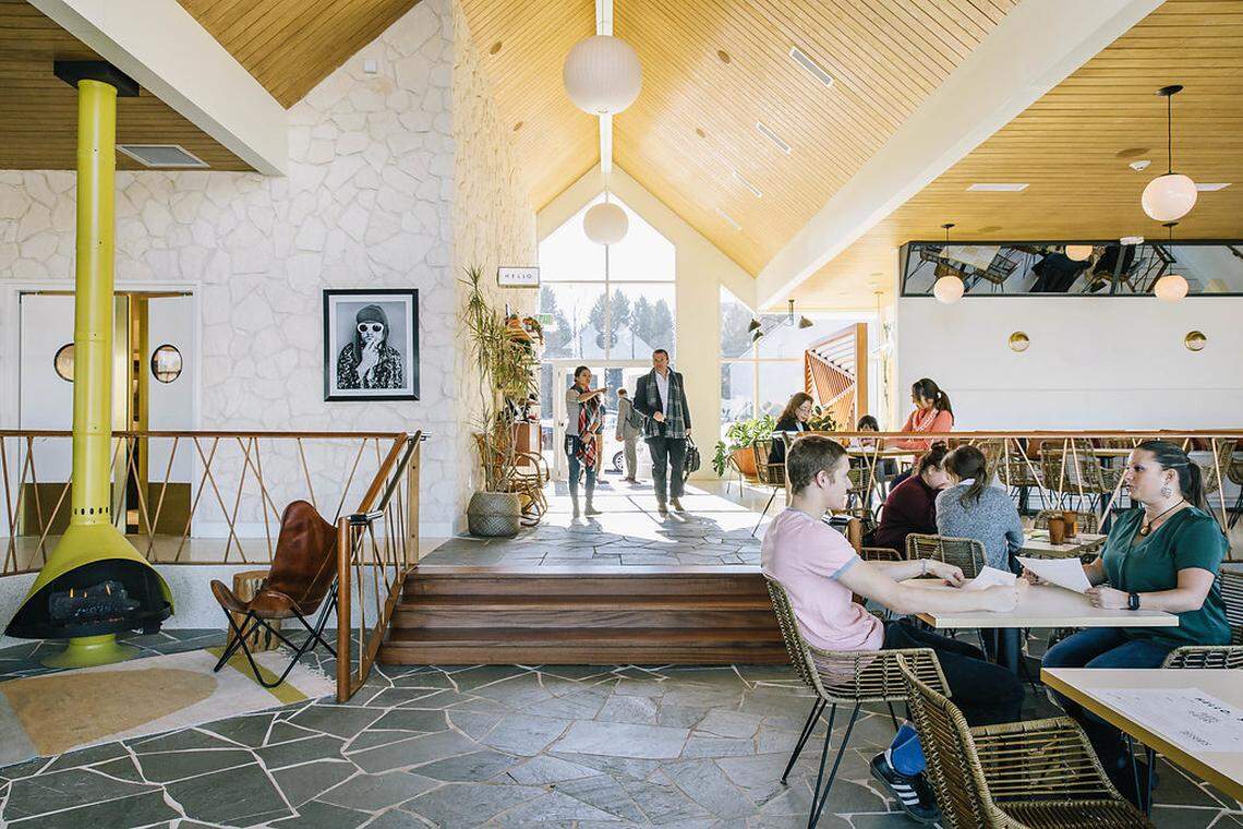A spacious and bright restaurant interior with a mid-century modern aesthetic. The room has a high, vaulted wooden ceiling with exposed beams and large windows at the far end, letting in natural light. A stone accent wall with a large framed picture of a man in sunglasses and a hat is prominent on the left. In the foreground, a couple is seated at a table, looking at papers. Behind them, other patrons are seated at tables in a slightly elevated dining area. On the left, a unique yellow freestanding fireplace sits on a mosaic-tiled floor. The space features a mix of textures and materials, including stone, wood, and metal.