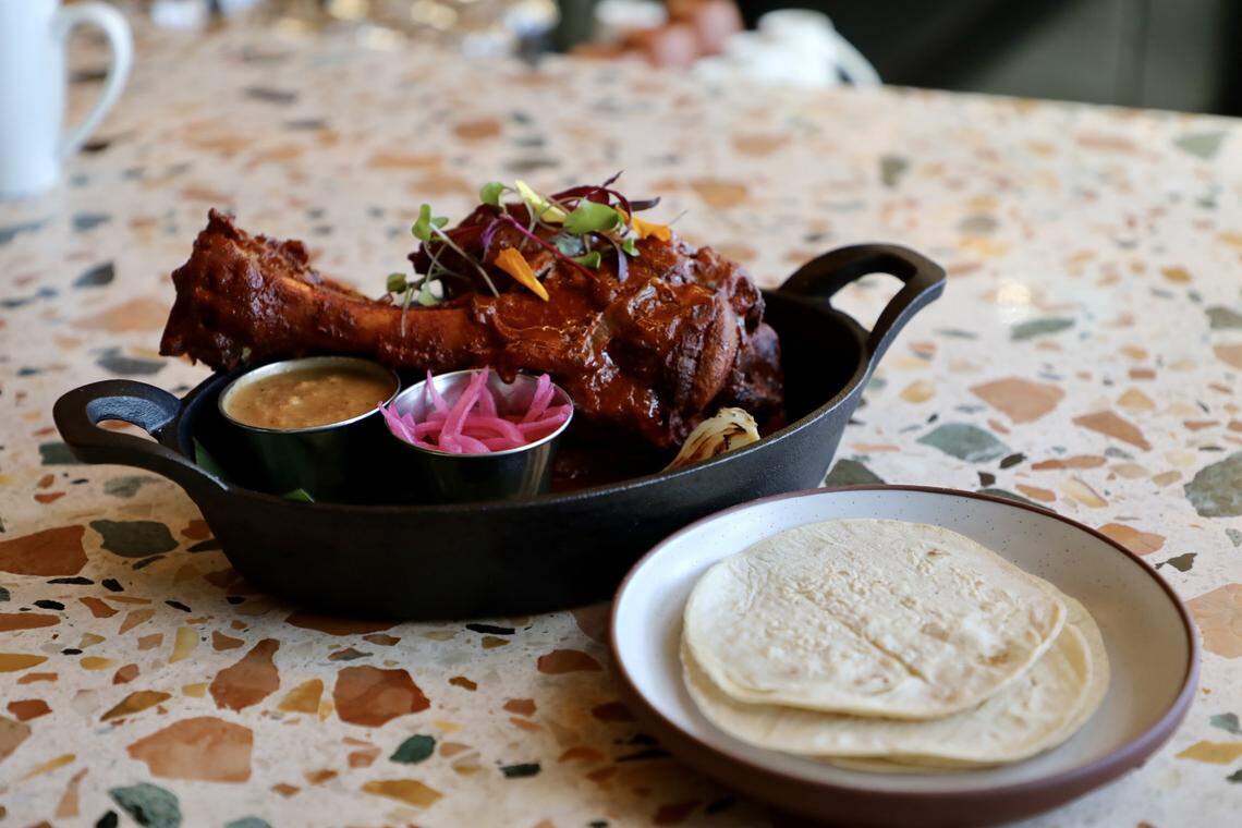 A medium-angle, slightly side-view shot shows a serving of Cochinita Pibil presented in a small, black cast-iron server with two handles, resting on a multi-colored terrazzo tabletop.