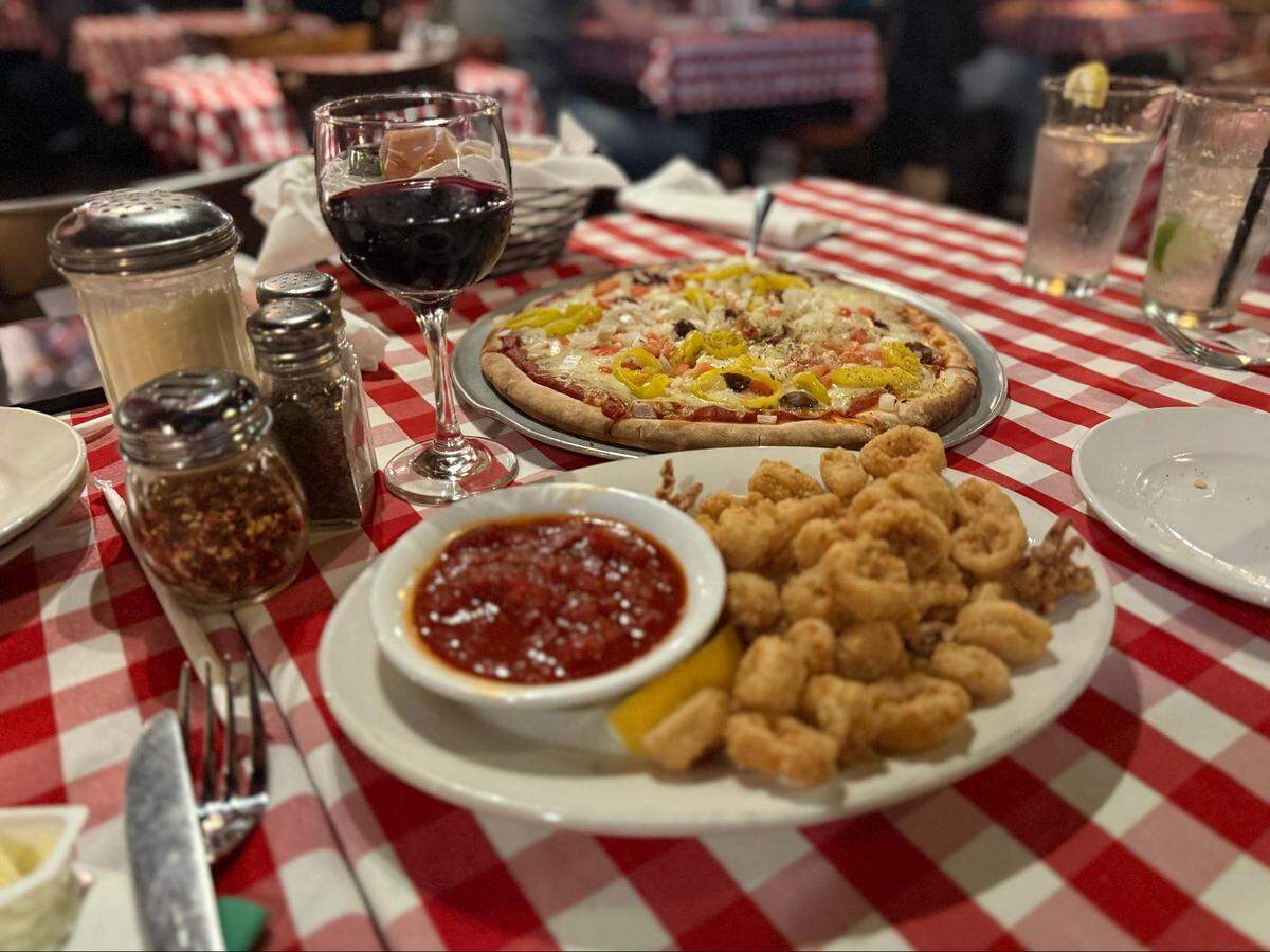 A warm and inviting scene at an Italian-American restaurant, with a red and white checkered tablecloth covering the table. In the foreground, a white plate holds a mound of golden-brown fried calamari next to a small white bowl of red marinara sauce, garnished with a lemon wedge. A wine glass filled with red wine sits prominently. Behind it, a whole pizza topped with cheese, olives, and yellow banana peppers is on a metal pan. Various tabletop items are scattered around, including shakers for grated cheese and red pepper flakes, salt and pepper shakers, and rolled silverware. In the background, other tables with similar tablecloths and blurred patrons create a lively atmosphere. Two tall glasses with clear drinks are visible on the right.