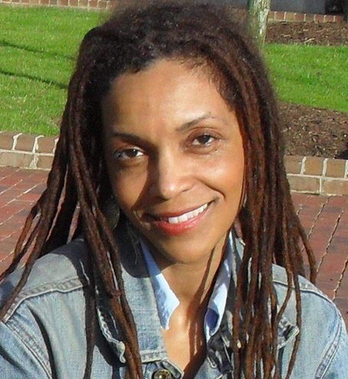 A close-up, head-and-shoulders shot of a woman with long brown dreadlocks, smiling warmly at the camera. She is wearing a light blue collared shirt under a denim jacket, and the outdoor background appears to be a brick path next to a lawn.