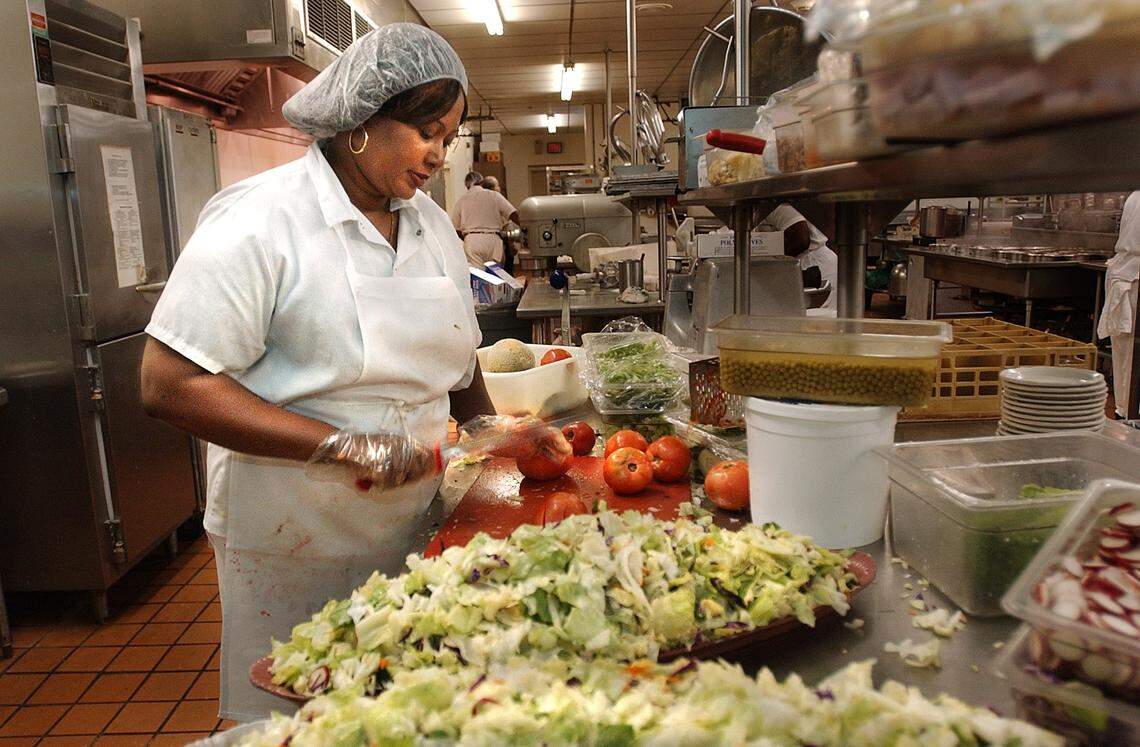 A cook in a professional kitchen wearing a white uniform, white apron, and a hair net is standing at a stainless steel prep counter, slicing tomatoes. A large pile of shredded lettuce and other cut vegetables is on the counter in the foreground.