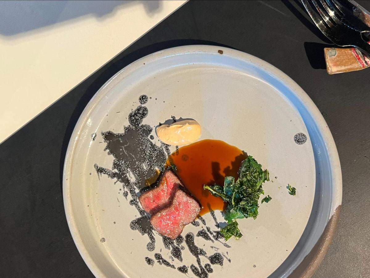 An overhead, angled shot of a meticulously plated dish featuring two thin slices of rare red meat. The meat is arranged next to a small pool of dark sauce and a cluster of green leafy vegetables. A dollop of aioli and abstract black splatters complete the presentation on the matte gray plate.