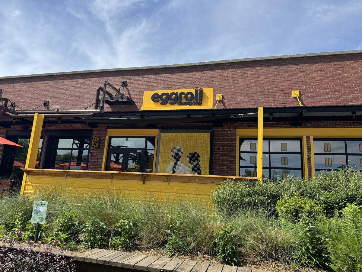 A brick building exterior featuring the “Eggroll Company” storefront. The shop is highlighted by a bright yellow sign and a matching yellow wooden patio barrier. Large windows show an interior with yellow walls and a “Big Dreams Start Small” graphic. Lush green ornamental grasses and a small “Native Plant Habitat” sign are in the foreground under a bright, clear blue sky.
