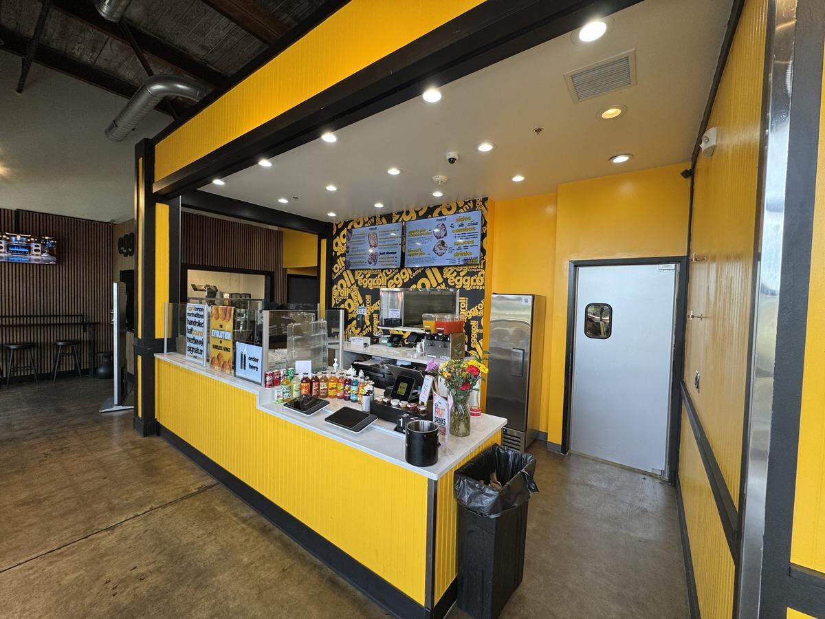The interior of Eggroll Company, featuring a bold yellow and black color scheme. A white service counter holds digital ordering tablets, various bottled sauces, and a vase of flowers. Behind the counter, digital menu boards display “Biscoff Apple Pie” and other specialty eggrolls against a wall covered in “Eggroll Company” typography wallpaper.