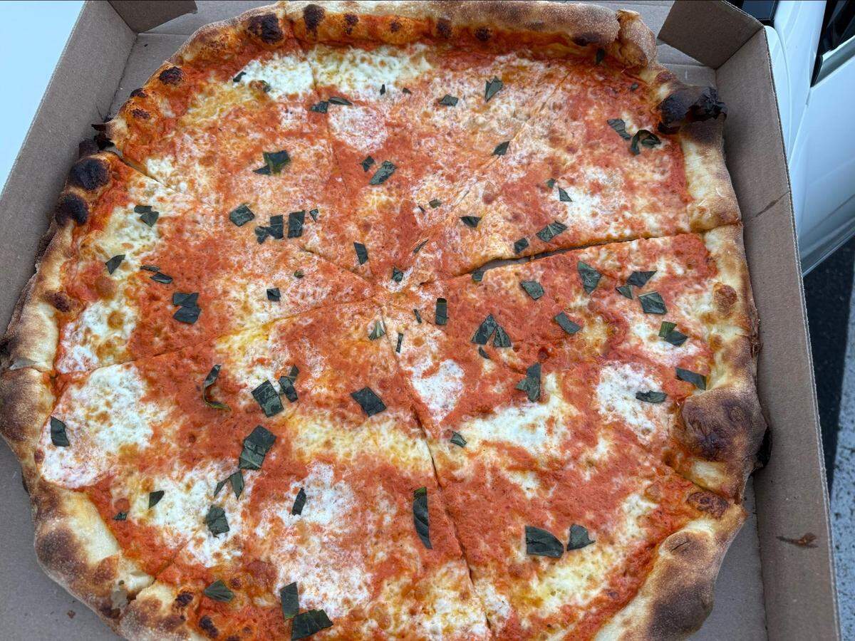 An overhead, close-up shot of a large, New York-style pizza inside an open cardboard box. The pizza is topped with red tomato sauce, dollops of melted mozzarella cheese, and chopped basil, and features a browned, bubbled crust.