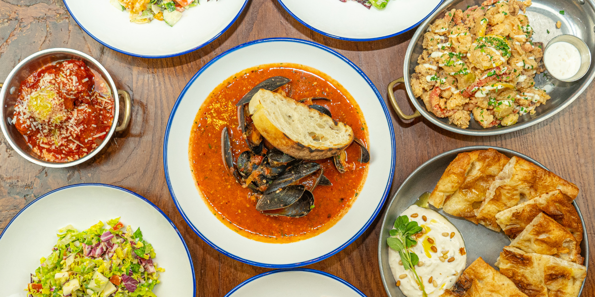 An overhead flat lay of a variety of Italian dishes arranged on a wooden table. The central dish is a white bowl with a blue rim containing mussels in a tomato broth. Other visible dishes include meatballs in sauce, fried calamari, flatbread with a white dip, and several plates of fresh chopped salads.