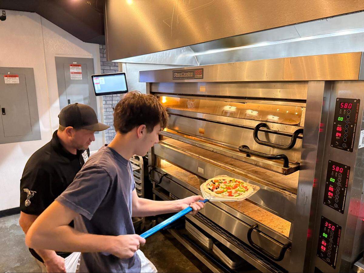 A young person uses a long-handled blue metal peel to slide a veggie-loaded pizza into a professional three-tier “PizzaMaster” deck oven. A chef stands beside him, monitoring the process. The oven’s digital displays show high cooking temperatures.