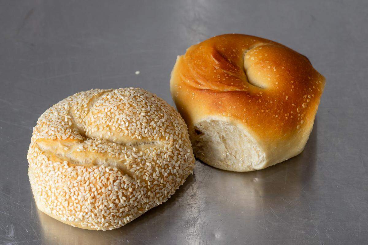 A close-up, eye-level shot of two round, golden-brown bagels on a shiny, metallic surface. The bagel on the left is covered with sesame seeds, while the one on the right is plain with a glossy, baked crust. Both have the characteristic swirled top.