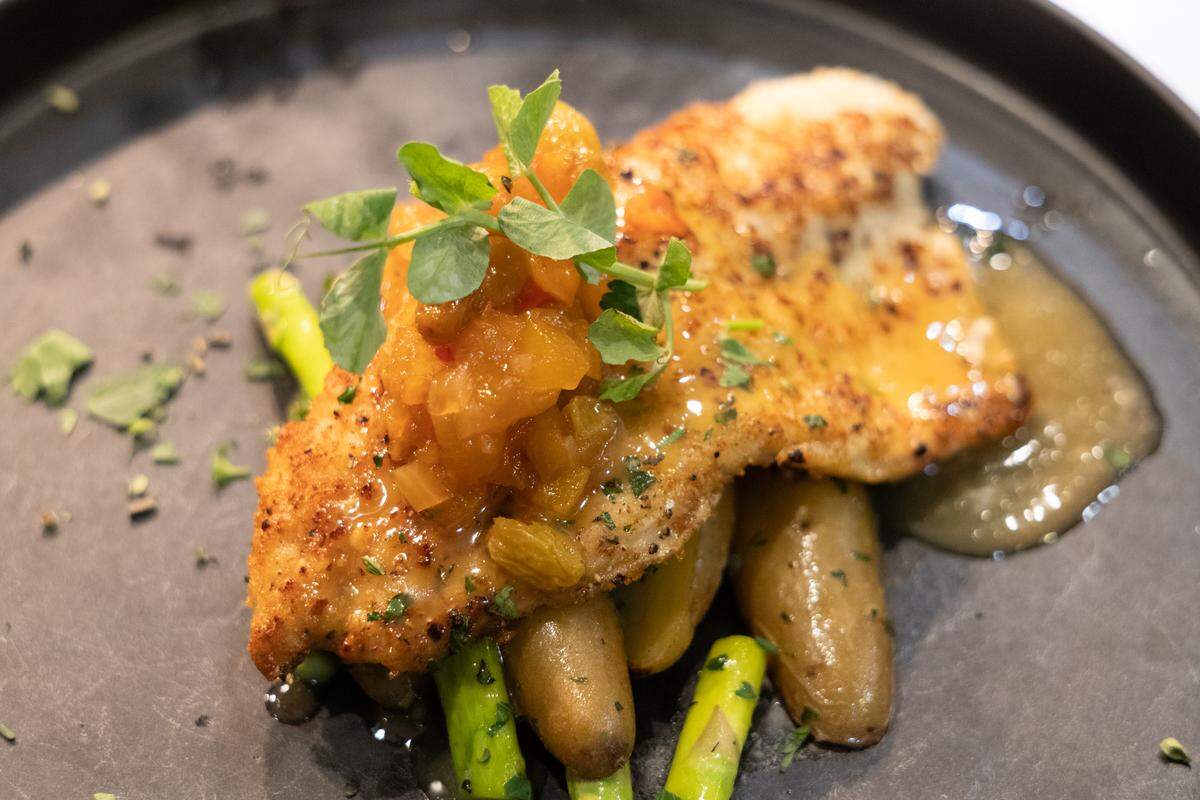A close-up, high-angle shot of a pan-seared white fish fillet served over roasted fingerling potatoes and bright green asparagus. The fish is topped with a vibrant fruit chutney and garnished with fresh pea shoots.