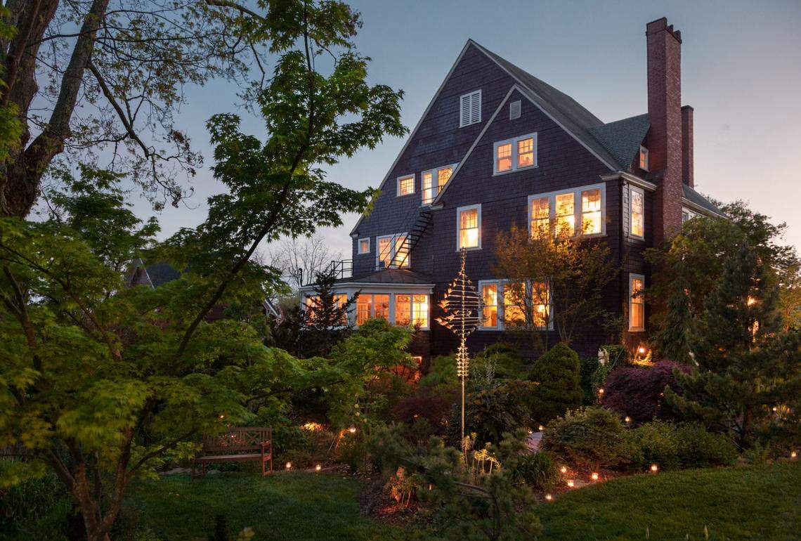 An exterior photo taken at twilight of the Blind Tiger Asheville guesthouse, a large, dark shingle-style house with many illuminated windows, a steep pitched roof, and a prominent brick chimney. The foreground features a lush, manicured garden with a wooden bench, lit by numerous small ground lights and flanked by large trees.