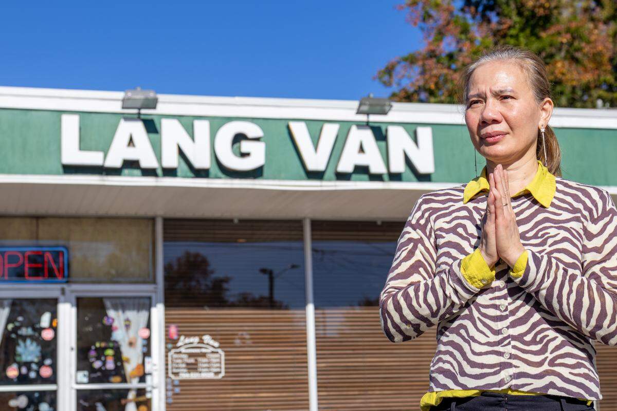 A medium, slightly low-angle shot of a woman with her hands pressed together in a grateful gesture, standing in front of a restaurant. She is wearing a zebra-print cardigan. In the background, the restaurant’s large green sign with white letters reading “LANG VAN” is prominent, along with a neon “OPEN” sign in the window, all set against a clear blue sky.