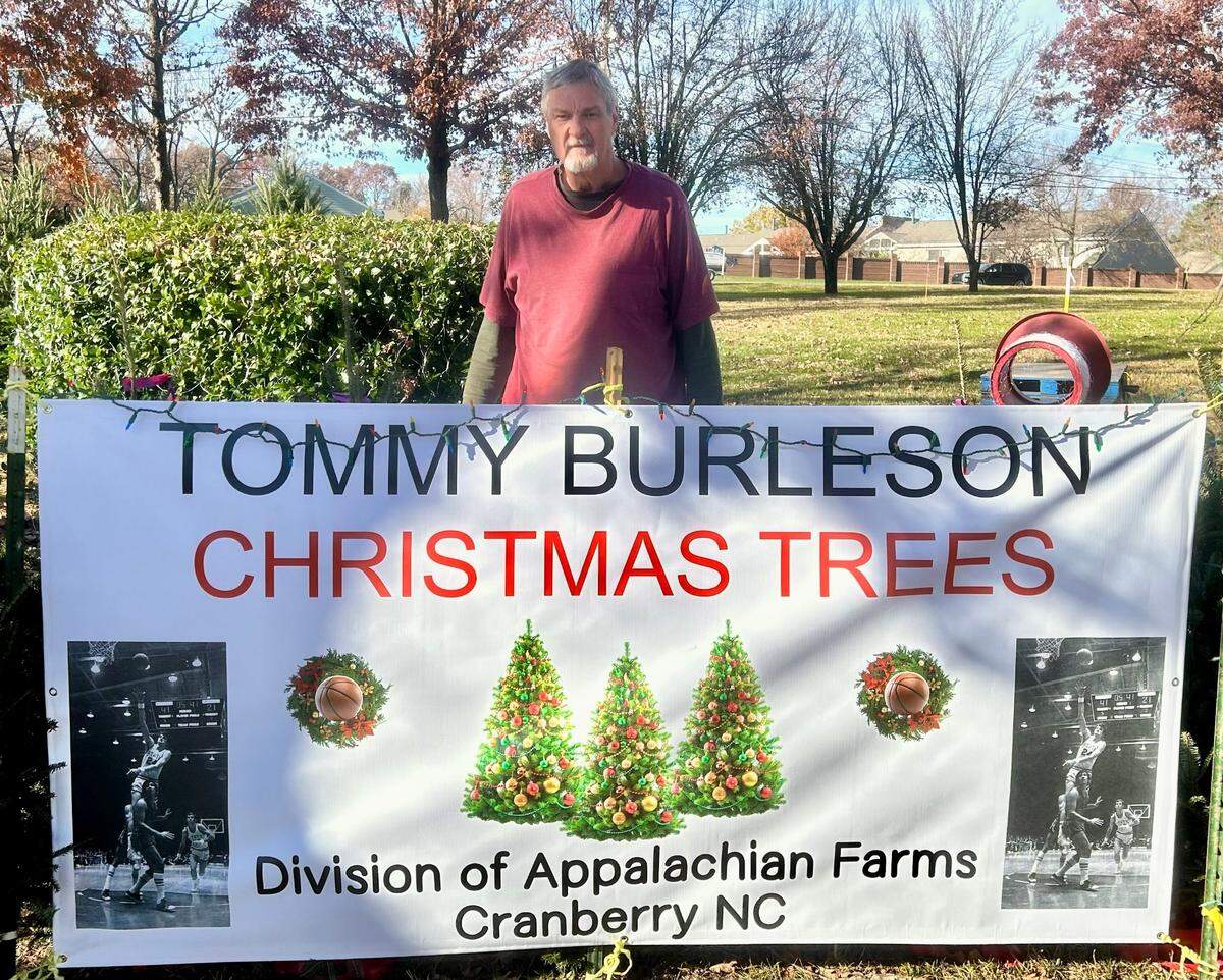 Tommy Burleson, the former N.C. State basketball player and member of the 1974 Wolfpack team that won the national title, stands behind the sign advertising his Christmas tree lot in Huntersville at Lake Norman Baptist Church. Burleson, who is from Avery County in the N.C. mountains, said he is donating his profits from the the sale of trees of Hurricane Helene relief.
