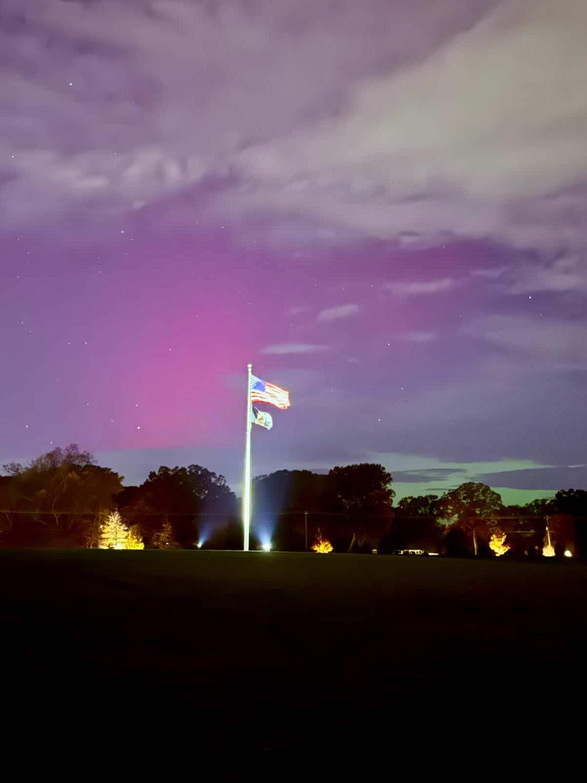 A low-angle nighttime photo capturing a purple and pink aurora borealis in a cloudy sky above a field. In the center, a brightly illuminated flagpole stands tall, flying the American and North Carolina flags. The horizon is marked by a dark line of trees with several bright, ground-level spotlights illuminating them from below, highlighting the extraordinary color of the sky.