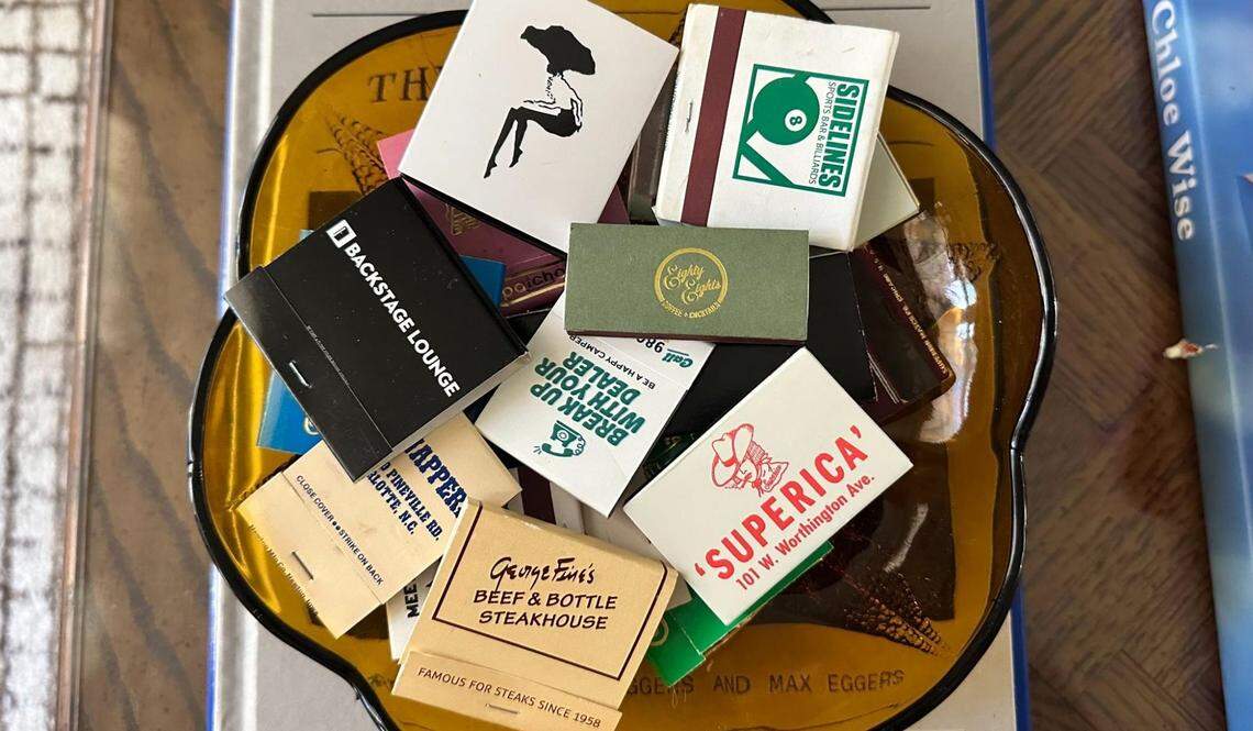 A close-up, overhead shot of a yellow, flower-shaped glass dish brimming with a collection of souvenir matchbooks from various restaurants and bars. Prominently visible are matchbooks for “BACKSTAGE LOUNGE,” “SUPERICA,” “George & Enid’s BEEF & BOTTLE STEAKHOUSE,” and “SIDELINES.” The dish is resting on a book on a coffee table.