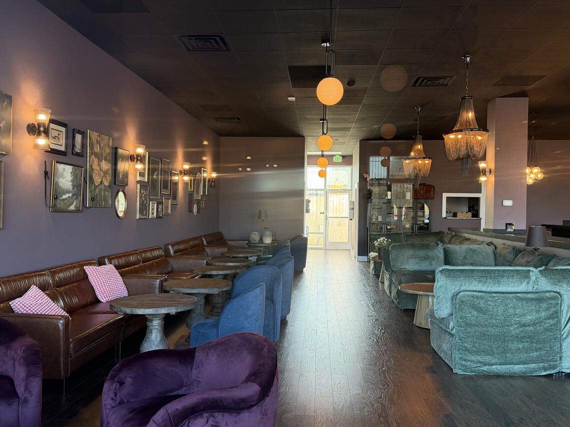 A cozy lounge area inside the wine bar with a moody, sophisticated atmosphere. Along the left, a plum-colored wall is decorated with a gallery of vintage-style framed paintings and lit by warm wall sconces. Seating includes long brown leather sofas, teal velvet armchairs, and deep purple velvet tub chairs arranged around small, round stone tables. Ornate beaded chandeliers and amber globe lights hang from the dark ceiling, creating a warm, intimate glow.