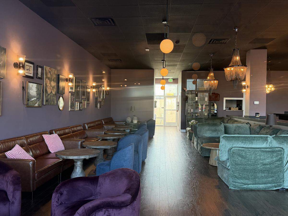 A cozy lounge area inside the wine bar with a moody, sophisticated atmosphere. Along the left, a plum-colored wall is decorated with a gallery of vintage-style framed paintings and lit by warm wall sconces. Seating includes long brown leather sofas, teal velvet armchairs, and deep purple velvet tub chairs arranged around small, round stone tables. Ornate beaded chandeliers and amber globe lights hang from the dark ceiling, creating a warm, intimate glow.