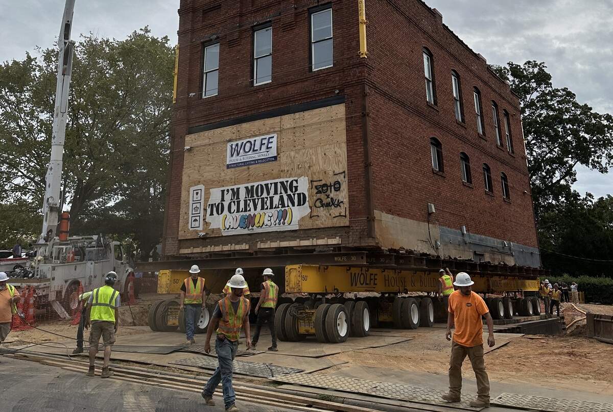 A wide shot of a historic red brick building being moved on a massive yellow hydraulic transport system by Wolfe House & Building Movers. A large banner on the side reads, “I’M MOVING TO CLEVELAND (AVENUE!!!)”. Several construction workers in high-visibility vests and hard hats are walking around the base of the structure as it maneuvers through a construction site.