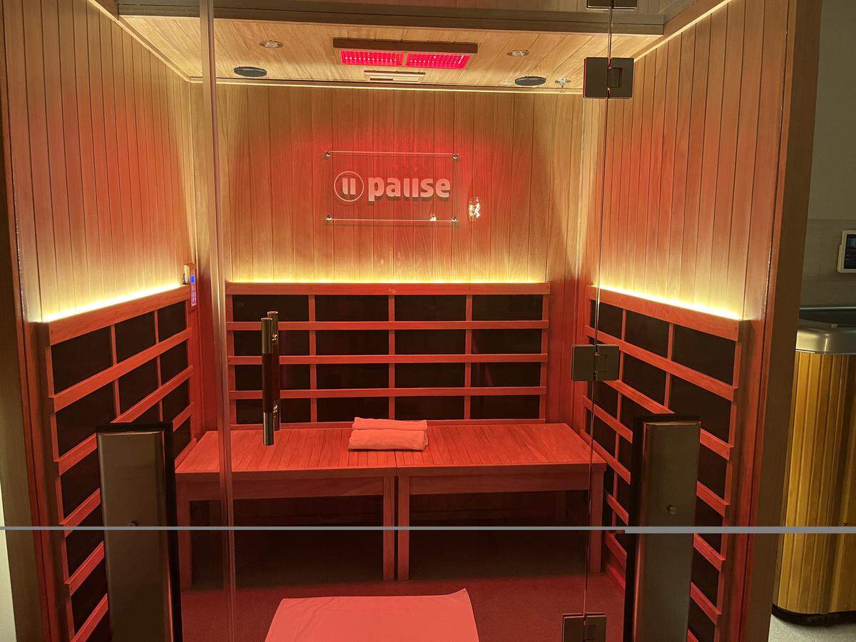 An eye-level, wide shot looking into a modern infrared sauna through a glass door. The interior features light-colored wood-paneled walls and a wooden bench with a white folded towel on top. Black heating panels are mounted along the lower walls, and the entire space is bathed in a warm red glow from an overhead light panel and a backlit logo on the back wall that reads “pause.” To the right, just outside the sauna, a portion of a wooden cold plunge tub is visible.