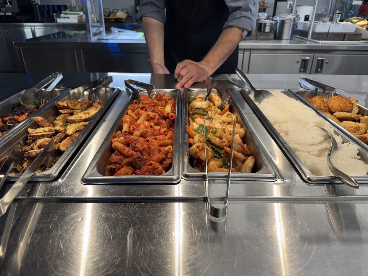 A close-up view of a stainless steel buffet line filled with five food trays. From left to right, the trays contain seasoned roasted potato wedges, rigatoni pasta in a red sauce, sautéed shrimp with green bell peppers, white rice, and breaded chicken cutlets. Metal serving tongs and spoons are placed in the trays, and the midsection of a chef in a dark apron is visible behind the counter.