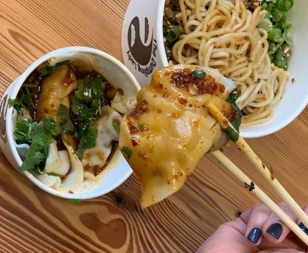 A person’s hand with dark-painted nails uses chopsticks to lift a dumpling from a white paper bowl. The dumpling is covered in a glossy red sauce and garnished with green onions. The bowl contains more dumplings in a dark sauce with fresh cilantro. In the blurred background, another white paper bowl, from “The Dumpling Lady,” contains a noodle dish with green onions and a dark sauce. The food is on a light wooden table.