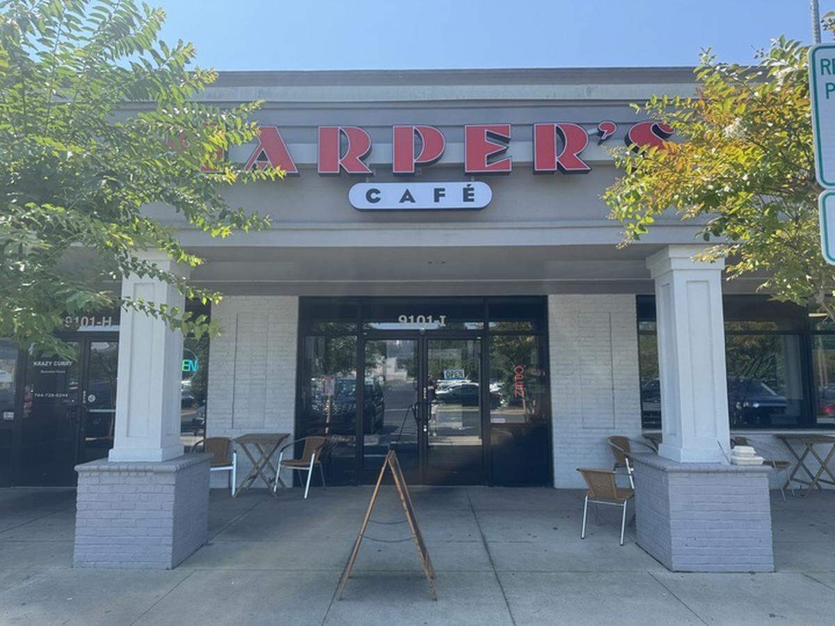 A frontal, medium-angle shot of the exterior of a restaurant called Harper’s Cafe. The restaurant is located in a one-story brick and stucco shopping center with large windows. The cafe’s sign is red with white lettering and is centered above the entrance. An A-frame sign sits on the sidewalk in front of the entrance, and there are two small outdoor tables with chairs on each side of the doorway.