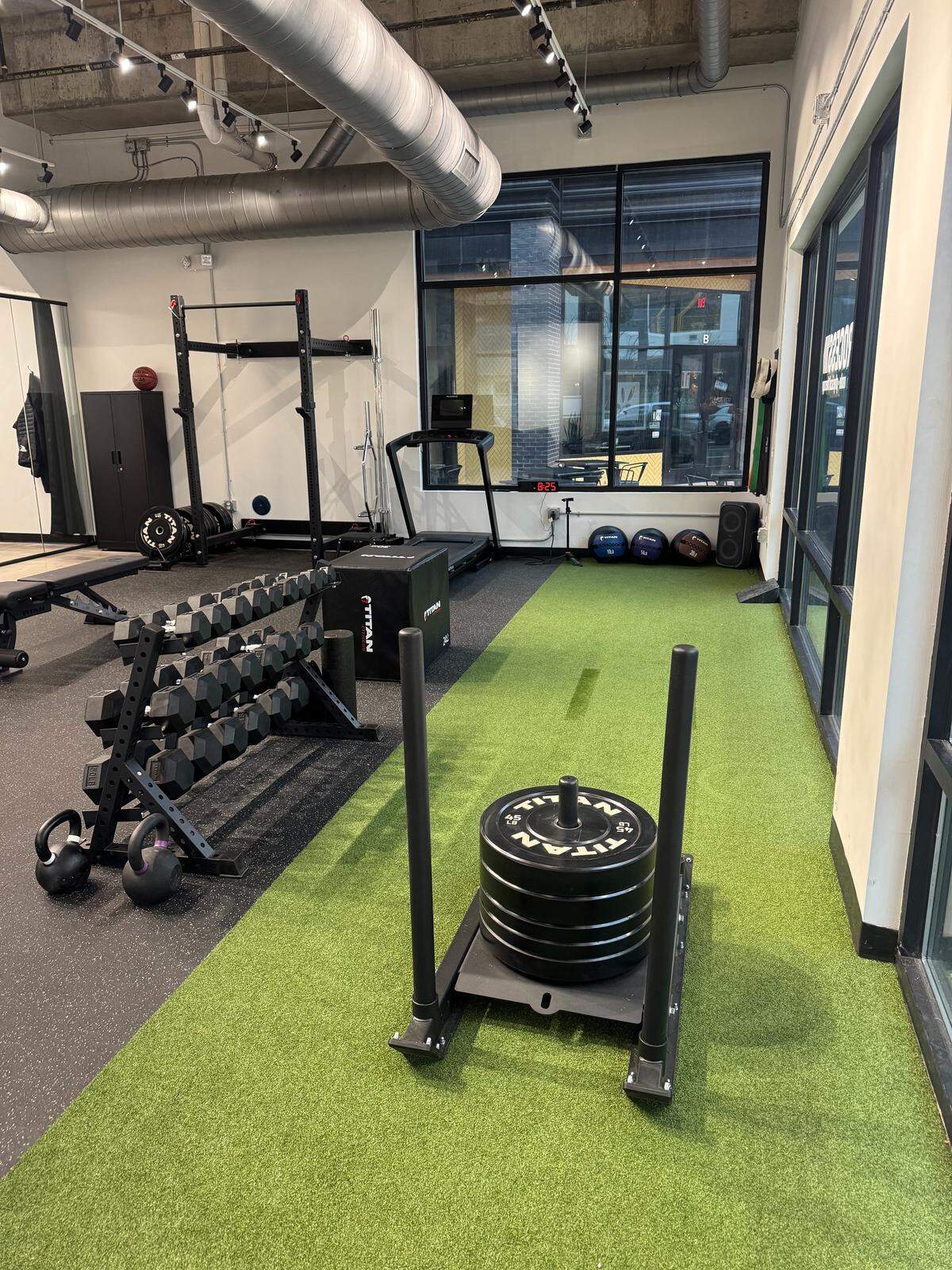 A functional training zone at Process Physical Therapy featuring a green turf lane with a weighted sled, a multi-tier dumbbell rack, and kettlebells. The industrial-style ceiling and track lighting create a modern, bright environment.