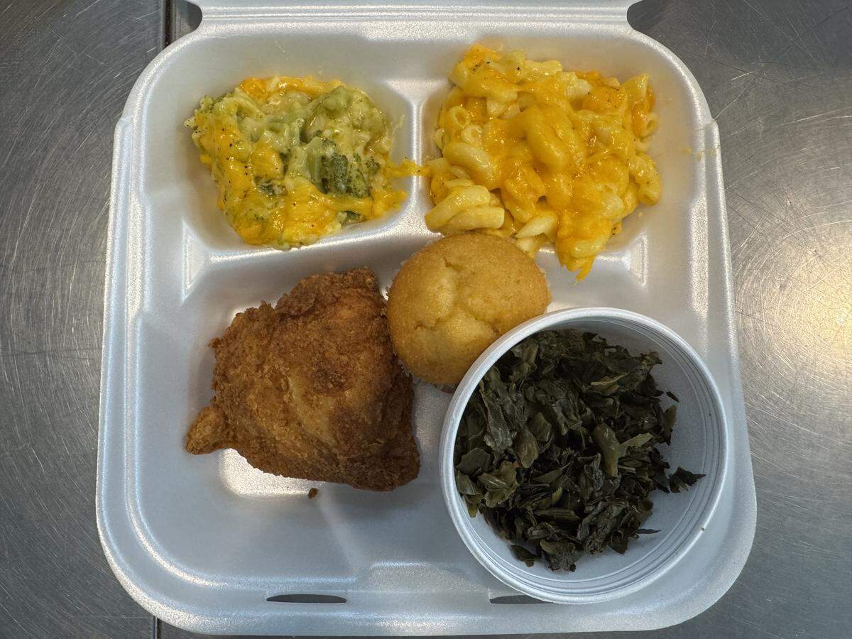 an overhead shot of a southern-style meal served in a white styrofoam three-compartment takeout container. The top left compartment holds broccoli cheese casserole, and the top right holds macaroni and cheese. The large bottom compartment contains a piece of crispy fried chicken, a round cornbread muffin, and a small white styrofoam cup filled with cooked collard greens.