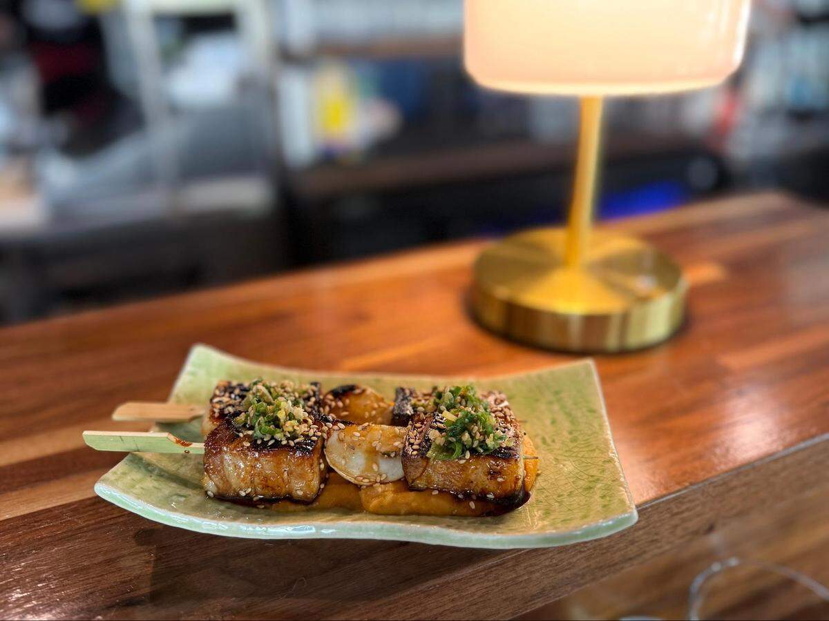 Two wooden skewers of grilled pork belly glazed in a dark, savory sauce. The meat is topped with sesame seeds and finely minced green scallions, served on a light green ceramic plate with a crackle glaze.