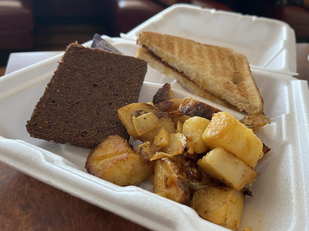 Food served in a white Styrofoam to-go container, featuring a large, thick slice of liver mush, a portion of home fried potatoes and onions, and a half-sandwich of toast.