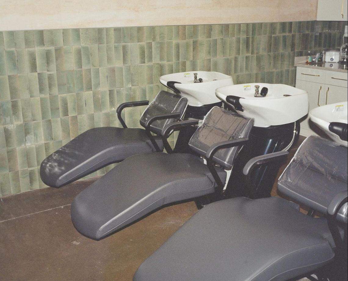 A row of three modern, black leather spa reclining chairs with attached white ceramic hair-washing basins. The background features a textured green tiled wall and a clean workstation.