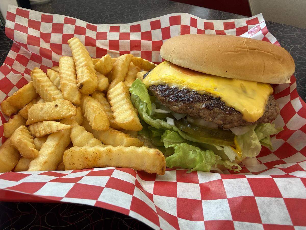 A close-up shot of a cheeseburger and a large pile of golden crinkle-cut fries served in a basket lined with red and white checkered paper. The burger features a toasted bun, a seasoned beef patty topped with melted yellow American cheese, fresh lettuce, and sliced pickles. The meal is set on a retro-style dark tabletop with a red-and-grey swirled pattern.