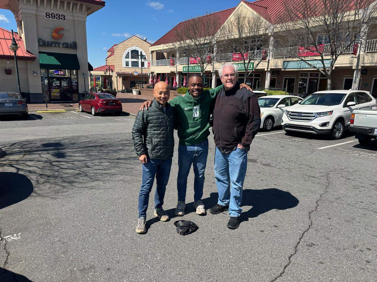 Three men stand smiling with their arms around each other in a sunny parking lot in front of a shopping center. The man on the left wears a dark quilted jacket and jeans; the man in the center wears a green hoodie and jeans; and the man on the right wears a black fleece jacket and jeans. In the background, storefronts include a “Crafty Crab” and other businesses under a clear blue sky.