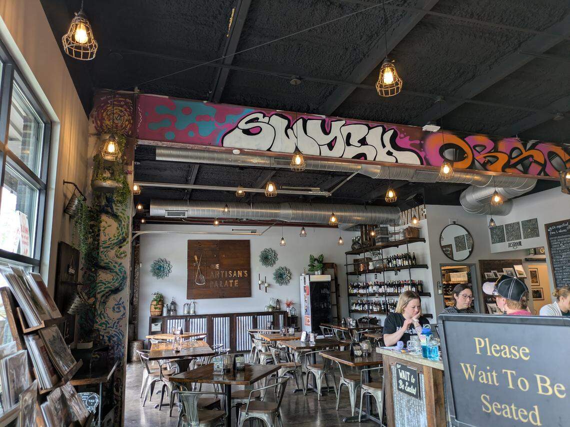 From the entrance, a view into The Artisan’s Palate restaurant and bar. The industrial-chic space is characterized by its exposed black ceiling and a large ceiling beam covered in colorful, stylized graffiti. The dining area has wood and metal furniture, and a large sign with the restaurant’s name hangs on the far wall.