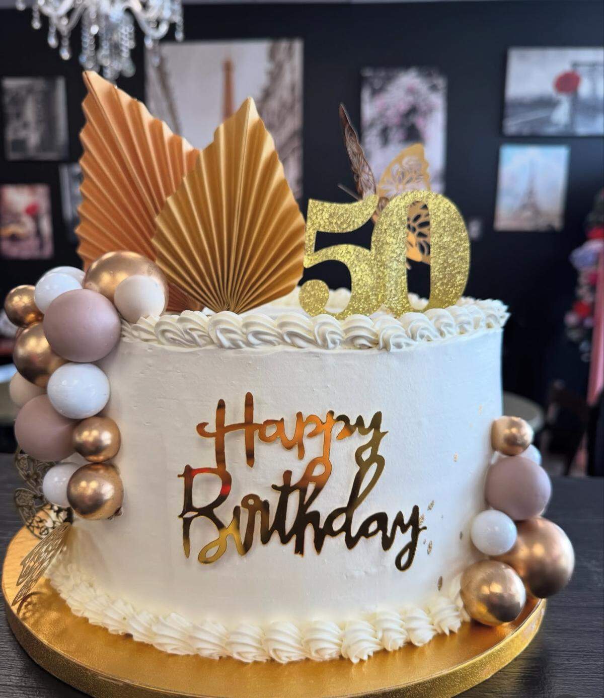 A beautiful white birthday cake is shown on a gold cake stand. The cake is decorated with a cascade of small balloons in shades of bronze, gold, and white. The top of the cake has white frosting swirls, two golden fan-shaped decorations, a gold glitter butterfly, and a gold glitter “50” topper. The side of the cake is decorated with a golden script that reads “Happy Birthday.” The background is a blurry wall with framed photos.