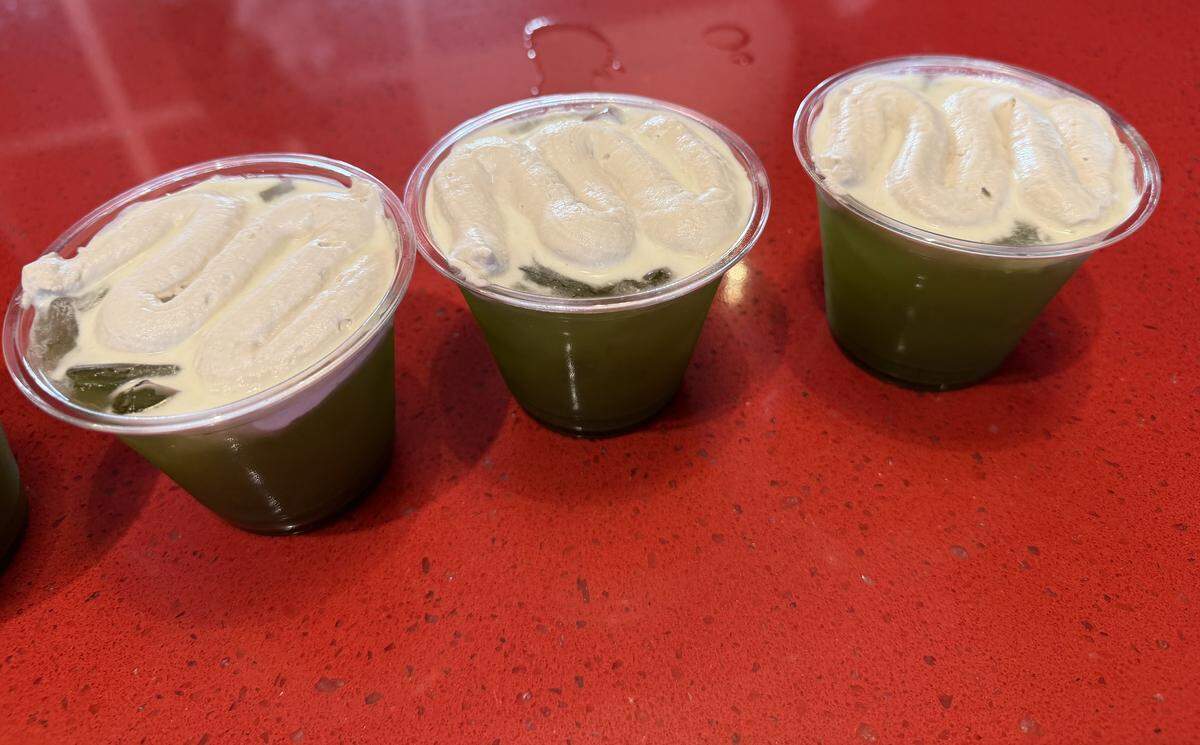 Three clear plastic cups filled with a vibrant green iced drink, each topped with a thick, wavy swirl of white foam. The drinks are lined up on a bright red speckled countertop.