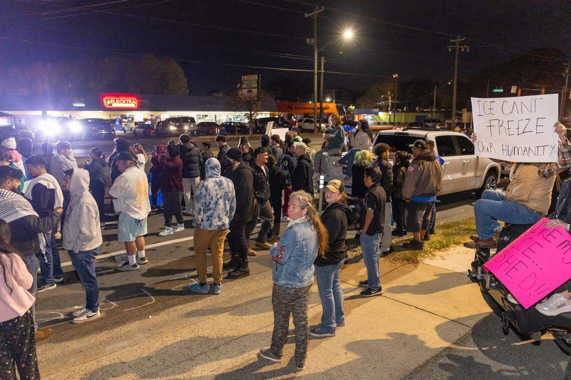 A long line of approximately 30-40 protesters stands on the edge of a street at night. They are facing a brightly lit row of storefronts, including one marked “MONACO NAIL SUPPLY.” Many protesters are holding white signs. The group is standing firmly on the asphalt near the sidewalk.
