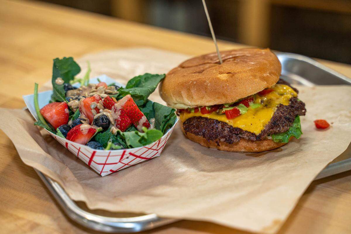 A cheeseburger and a side salad served on brown parchment paper atop a metal tray. The burger features a toasted bun, a grilled beef patty topped with melted yellow cheese, lettuce, and red and green sauces. Next to it, a small paper boat holds a fresh salad of kale or spinach mixed with strawberries, blueberries, and sunflower seeds, drizzled with a creamy dressing.