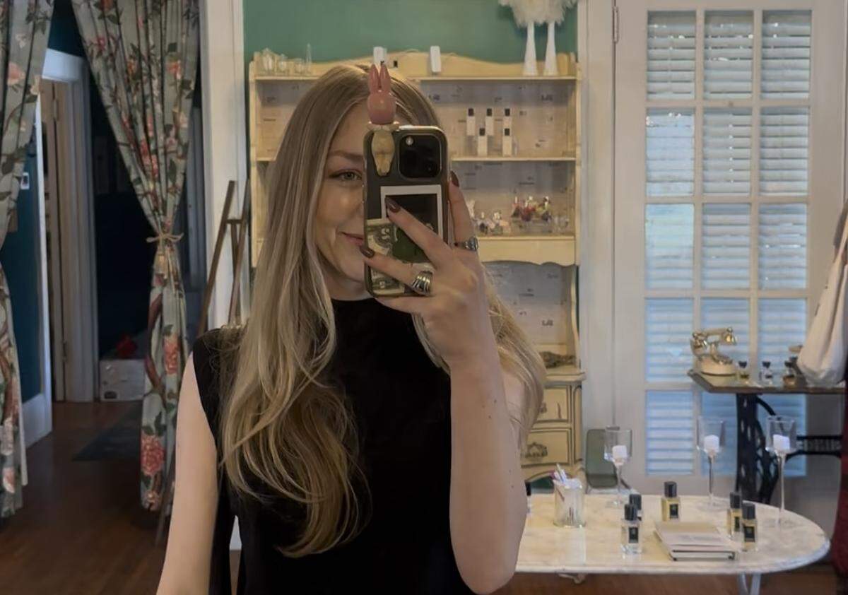 A mirror selfie of a person with long blonde hair and a black top,holding a phone with a rabbit-shaped accessory on top. The background shows the interior of the boutique, featuring a vintage cream-colored hutch filled with perfume bottles, a marble-top table, and floral patterned curtains.