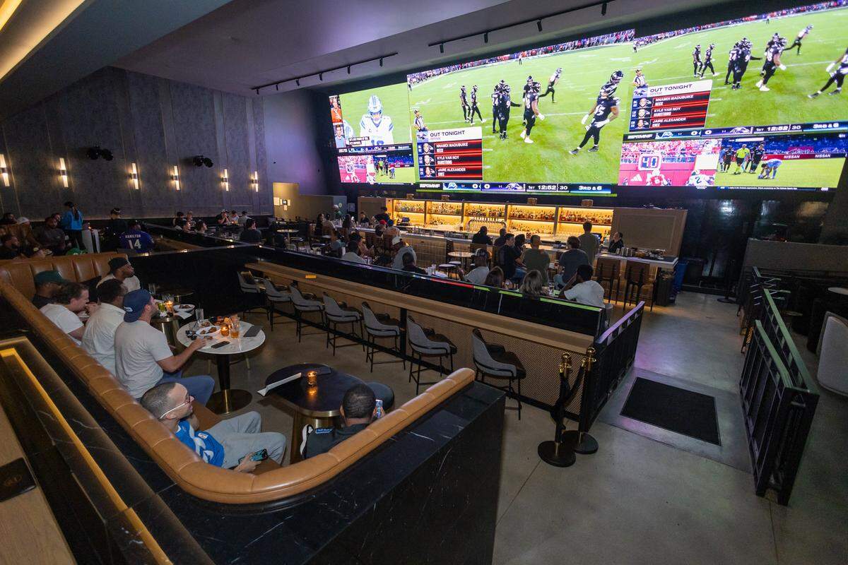 The interior of the Blinders sports bar is bustling with patrons watching a football game. A group of men in the foreground are seated in a large caramel-leather banquette, looking up at the huge video wall that spans the area above the main bar. The entire stylish, dimly lit space is focused on the live sporting event.