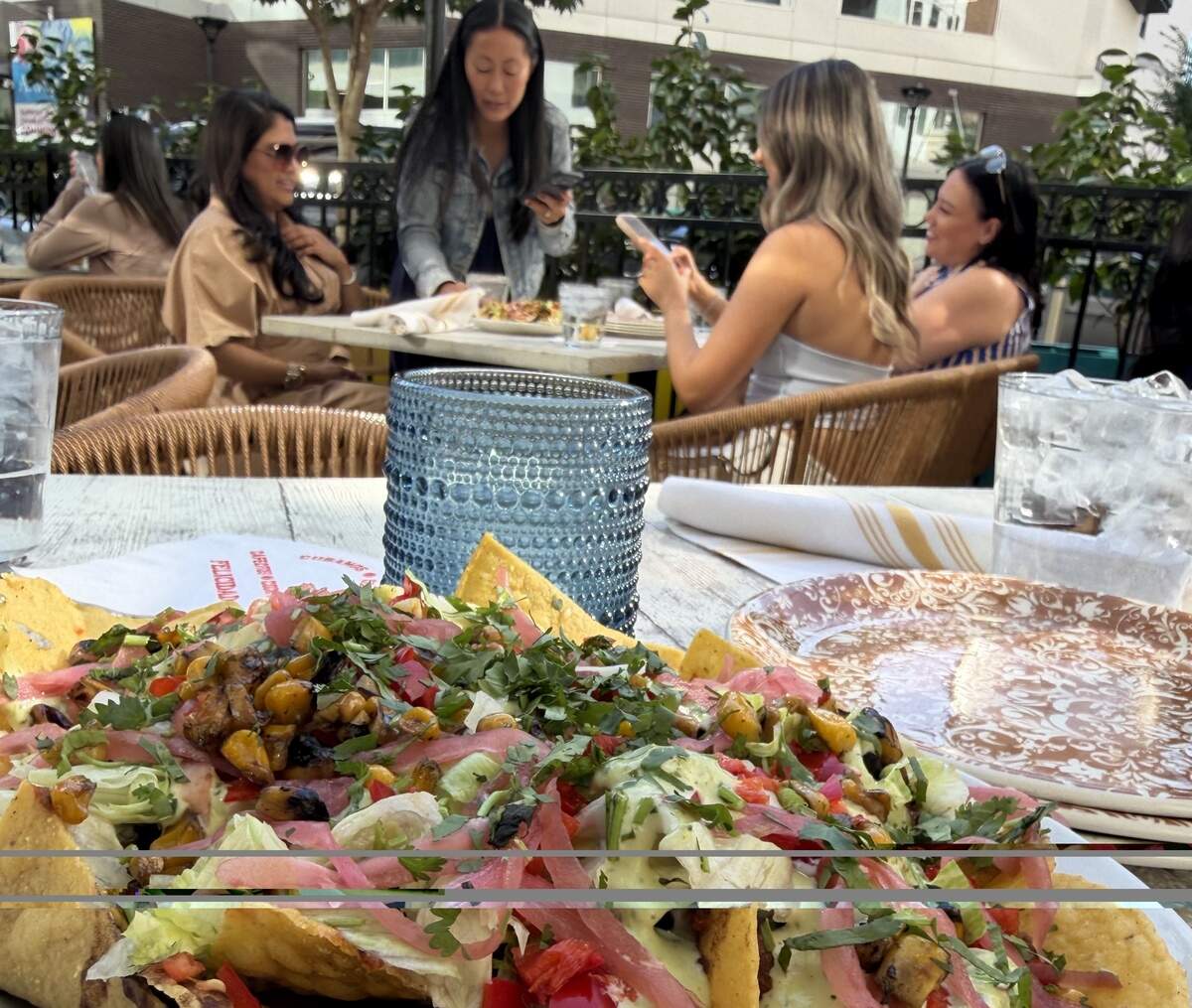 A close-up of a large plate of loaded nachos topped with roasted corn, pickled red onions, cilantro, lettuce, and crema on an outdoor restaurant table, with a blue textured glass, patterned plates, and diners blurred in the background.