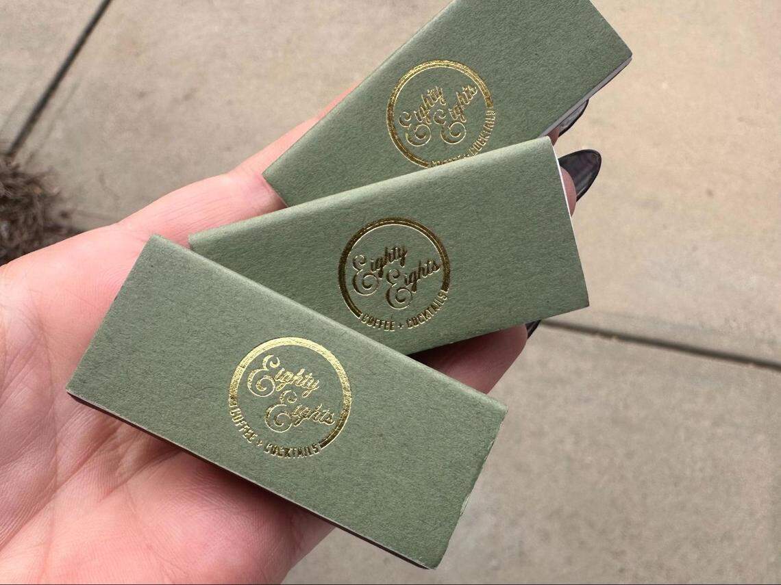 A close-up of a hand with dark nail polish holding three identical, sage green matchbooks. The matchbooks are fanned out to display a circular, gold foil logo on each, which reads “Eighty Eights” in a cursive font, with “COFFEE & COCKTAILS” underneath. The background appears to be a concrete sidewalk.