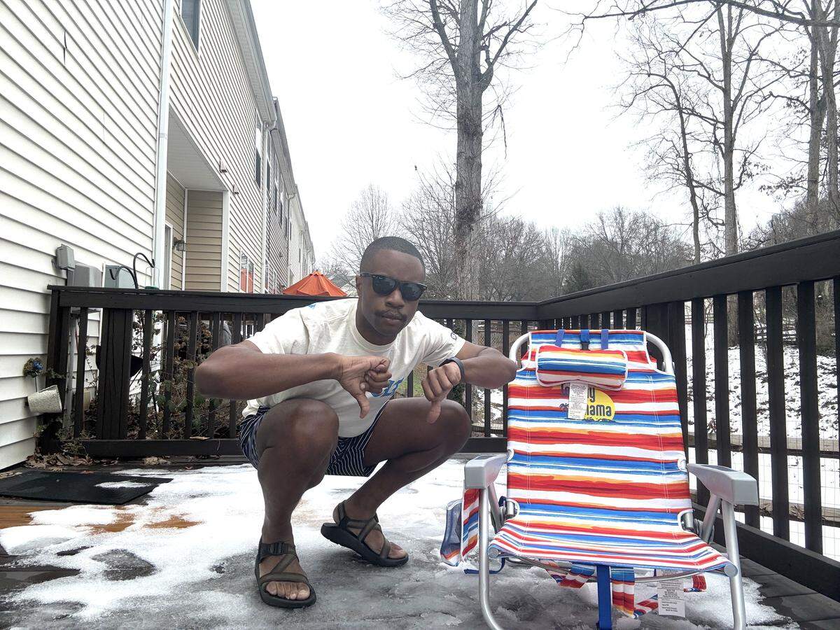 A man wearing sunglasses, a t-shirt, shorts, and sandals squats on a wooden deck partially covered in slushy snow, giving two thumbs down to the camera. Next to him sits a colorful, striped Tommy Bahama beach chair, highlighting the humorous contrast between his summer attire and the wintry weather.