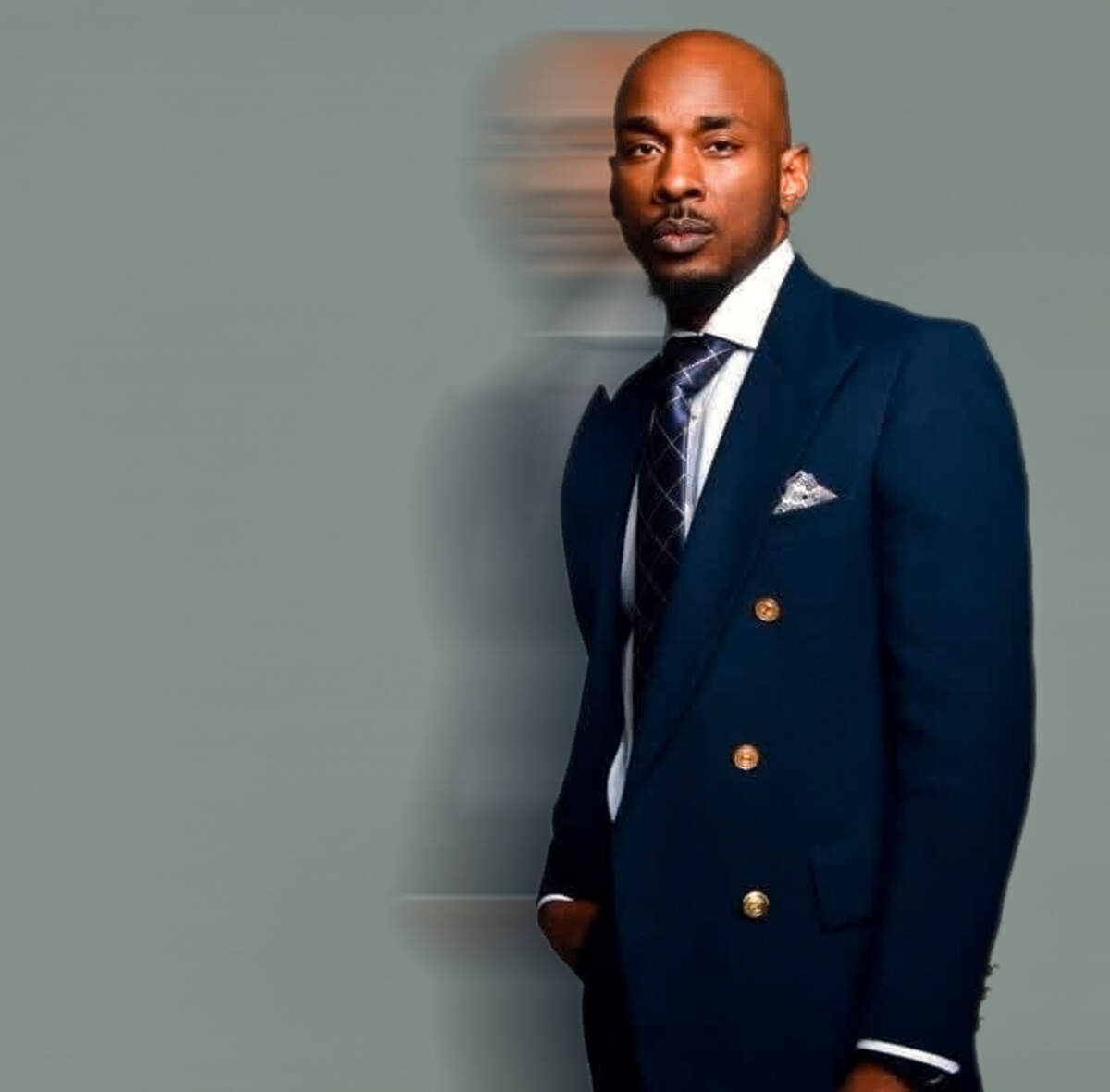 A bald man with a neat goatee stands in a three-quarter profile against a solid, muted gray-green background. He wears a sharp, double-breasted navy blue suit jacket with gold buttons, paired with a white collared shirt, a dark patterned tie, and a patterned pocket square. A stylized, translucent echo effect of his profile mimics his silhouette to the left, adding a dynamic layer to the portrait.