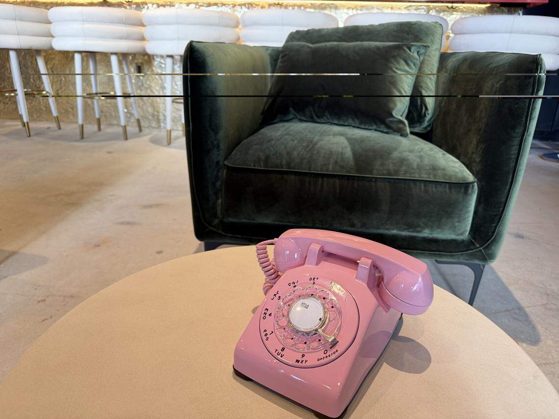 A vibrant pink vintage rotary telephone sits on a light beige round table in a stylish lounge. In the background, a plush dark green velvet armchair with matching pillows is positioned in front of a row of white modern bar stools with gold-tipped legs. The setting features a minimalist, chic aesthetic with industrial flooring.