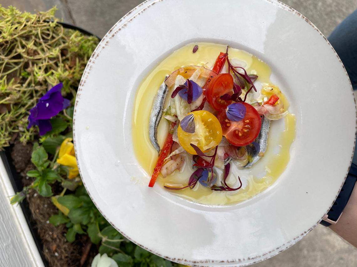 An overhead, close-up shot of a white appetizer plate featuring a dish of white anchovies. The anchovies are arranged with sliced red and yellow cherry tomatoes, slivers of red pepper, and small shrimp, all sitting in a pool of yellow olive oil and garnished with edible purple flowers. A person’s hand and a portion of a planter with yellow and purple flowers are visible on the left side of the frame.