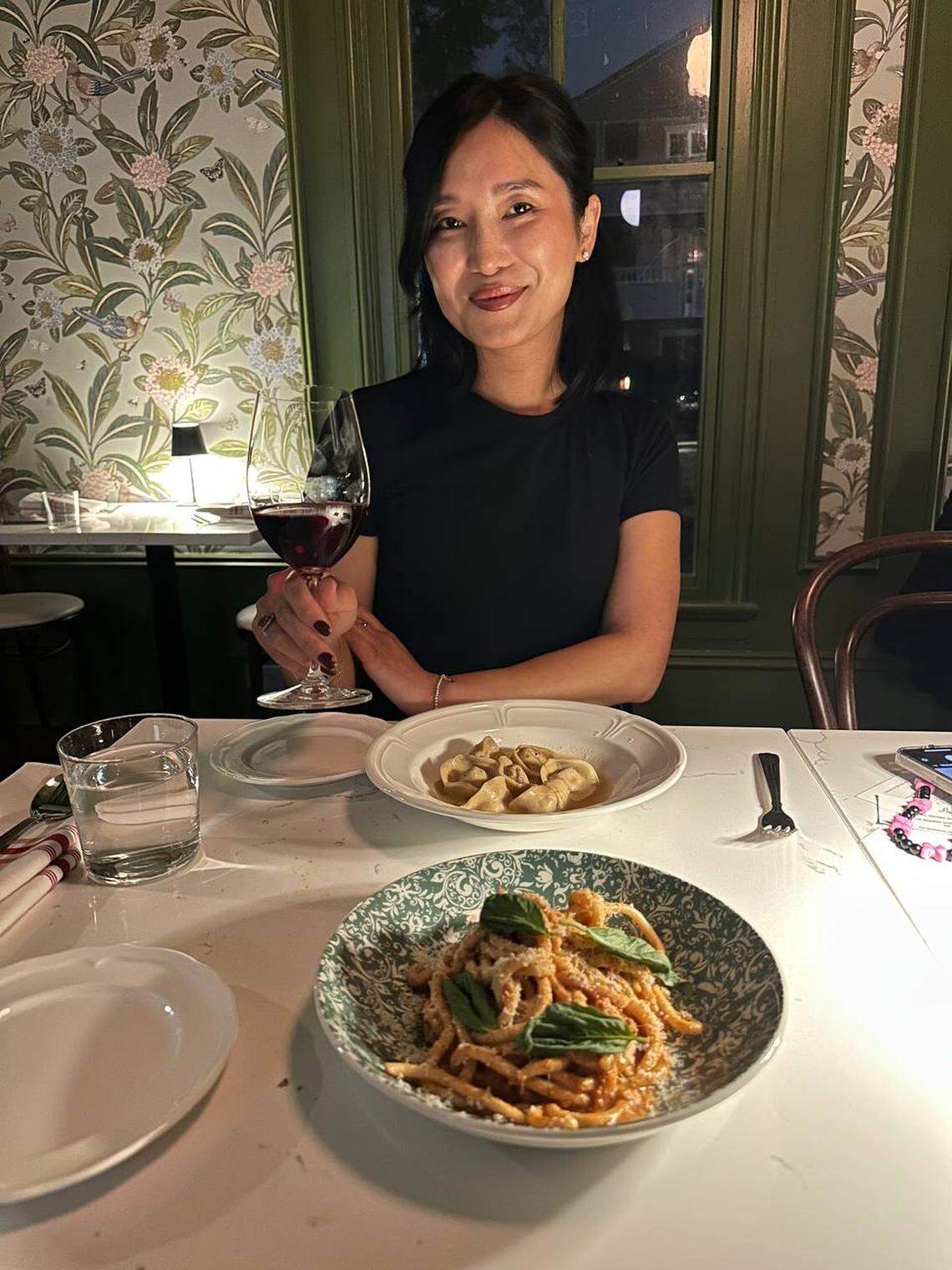 A person with short, dark hair sits at a restaurant table, holding a glass of red wine and smiling. In front of them are two dishes of pasta and white plates on a patterned tablecloth. The restaurant has green trim and wallpaper with a floral pattern.