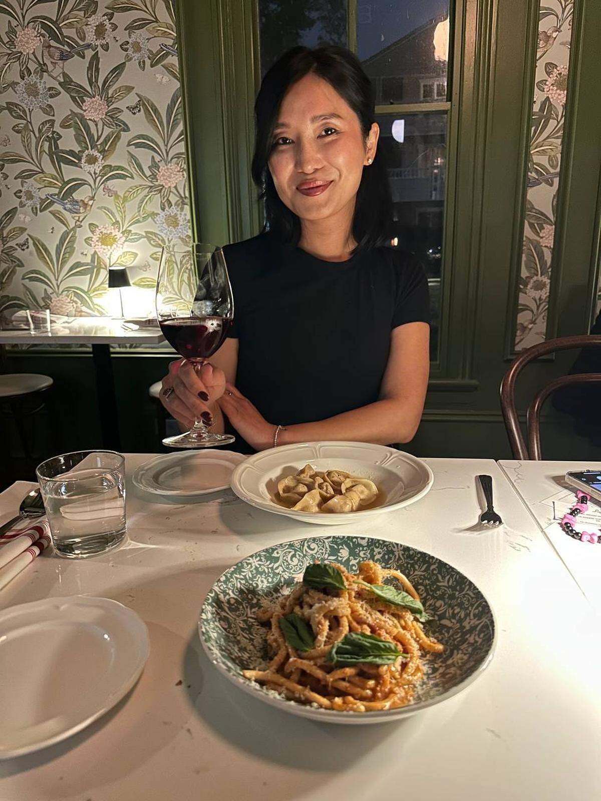 A person with short, dark hair sits at a restaurant table, holding a glass of red wine and smiling. In front of them are two dishes of pasta and white plates on a patterned tablecloth. The restaurant has green trim and wallpaper with a floral pattern.