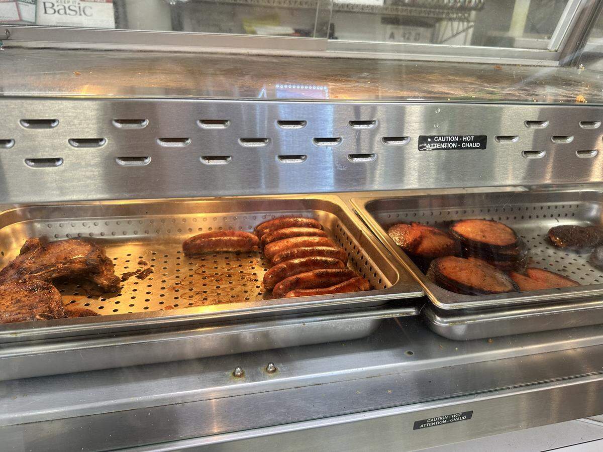 A close-up, eye-level shot of a stainless steel, self-serve hot food counter. The counter holds deep metal trays, one containing seasoned, grilled meat and a row of browned sausages, and an adjacent tray holding thick, round slices of pan-fried meat. “CAUTION - HOT” / “ATTENTION - CHAUD” warning labels are visible on the front of the unit.