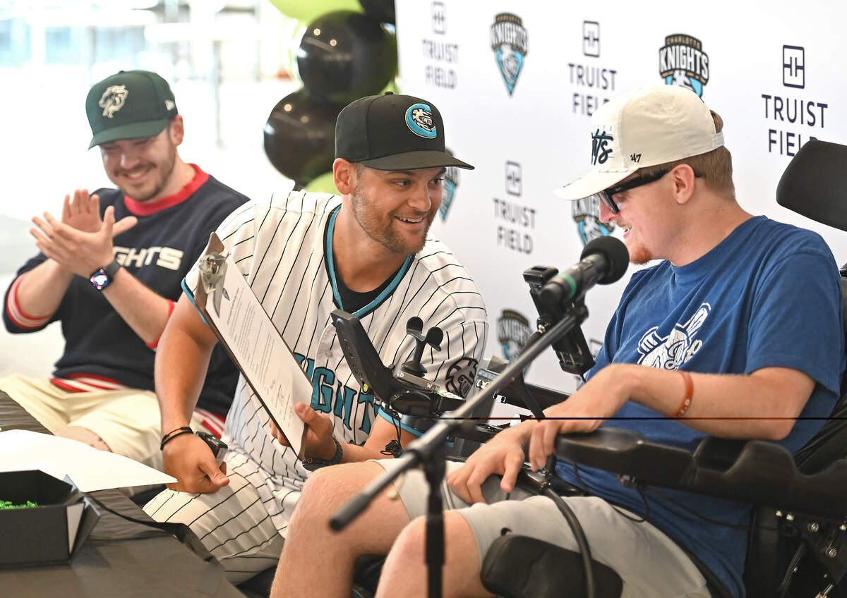 Charlotte Knights manager Chad Pinder, center, shows Nick Adair, right, the paper work for his one-day contract with the team during a press conference at Truist Field in Charlotte, NC on Saturday, June 27, 2026. Adair suffered a traumatic spinal cord injury that has left him paralyzed from the chest down. In addition to signing a one-day contract with the team, Adair designed along with Knights graphic designer Will Briggs a custom jersey that the team will wear during Saturday night’s game. The jersey features a glow-in-the-dark design.